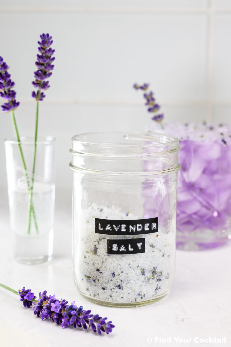 A glass jar labeled Lavender Salt sits on a white counter, surrounded by fresh lavender sprigs and a glass of lavender flowers in the background.
