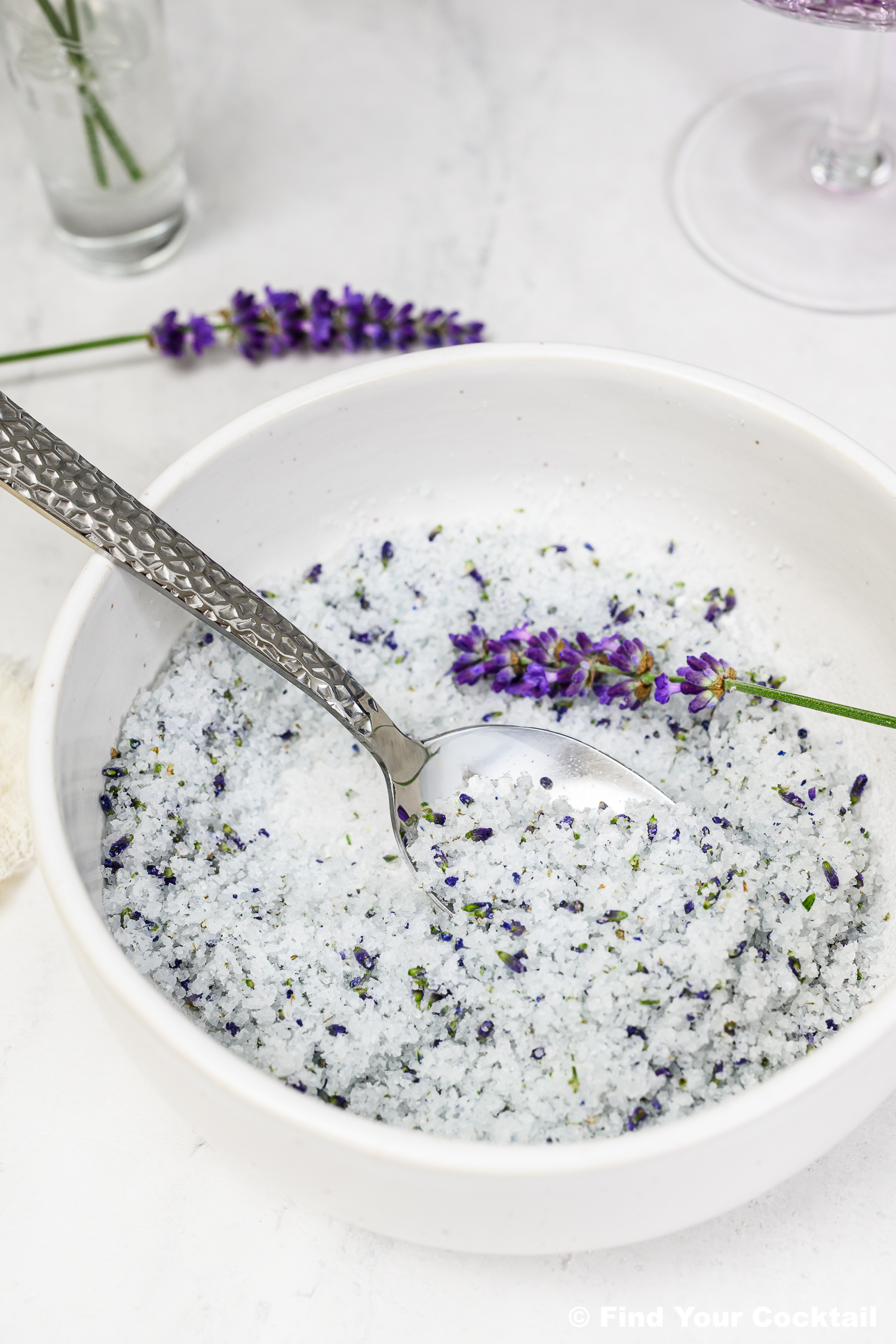 A white bowl filled with coarse salt mixed with dried lavender flowers, with a silver spoon and a lavender sprig on the rim.