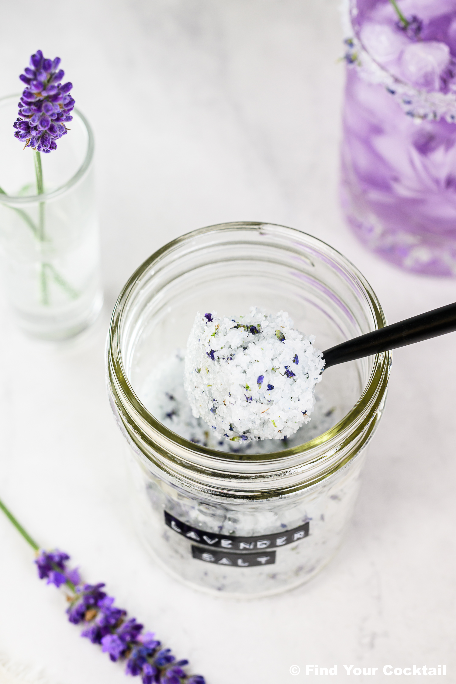 A spoon scoops lavender salt from a glass jar labeled lavender salt. Fresh lavender flowers are nearby on a white surface.
