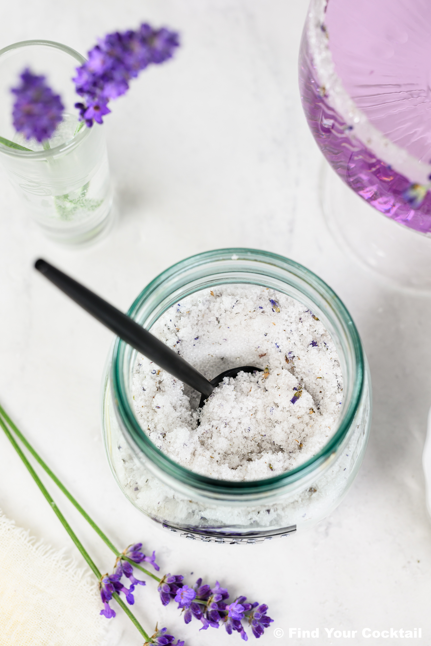 A jar of lavender sugar with a black spoon, fresh lavender sprigs, and a glass of purple liquid on a white surface.