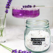 A jar labeled Lavender Sugar sits on a counter next to fresh lavender sprigs, a glass with a purple drink, and a small vase with lavender.