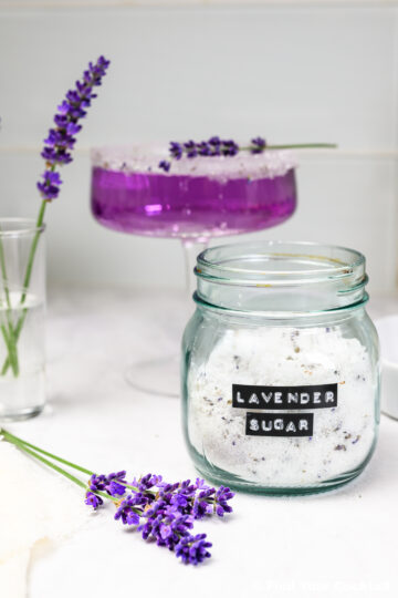A jar labeled Lavender Sugar sits on a counter next to fresh lavender sprigs, a glass with a purple drink, and a small vase with lavender.