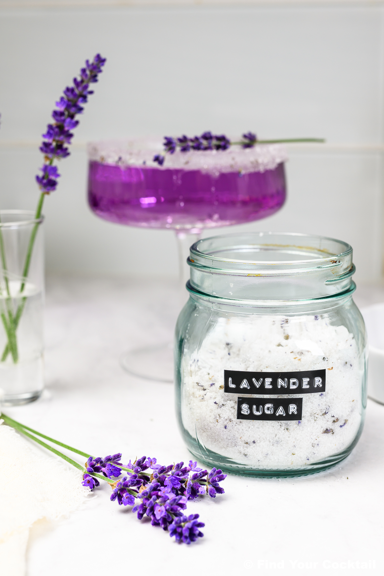 A jar labeled Lavender Sugar sits on a counter next to fresh lavender sprigs, a glass with a purple drink, and a small vase with lavender.