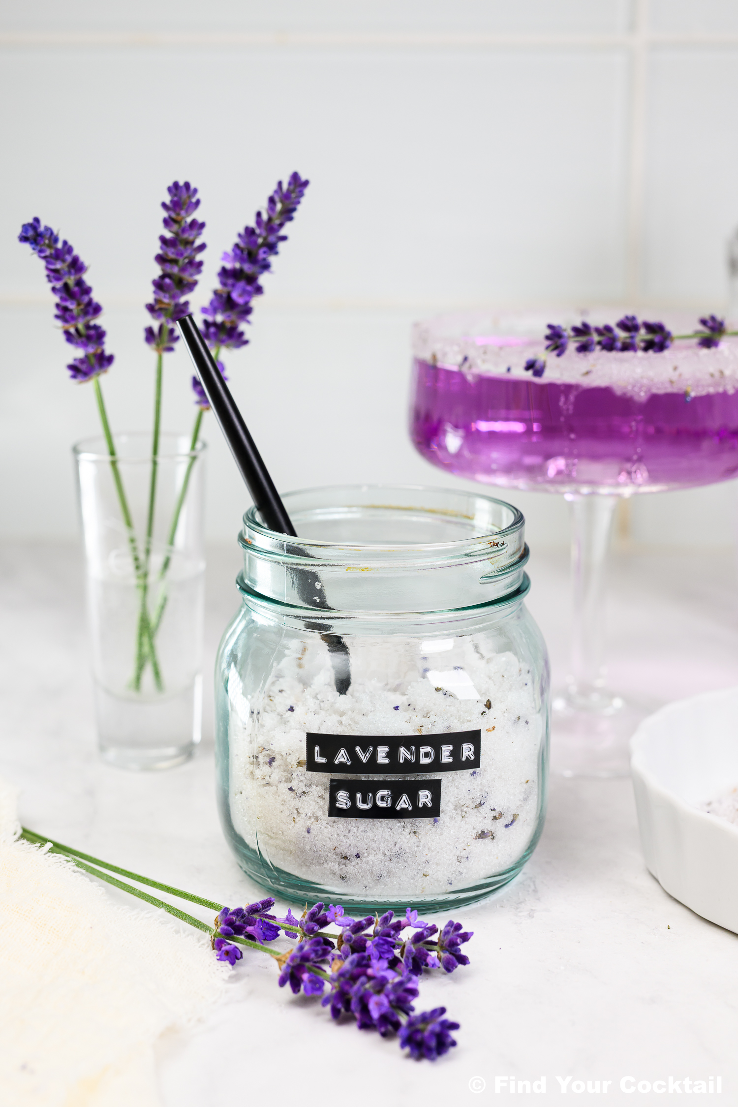 A glass jar labeled Lavender Sugar with a black spoon, fresh lavender sprigs, and a purple cocktail with a sugared rim on a white counter.