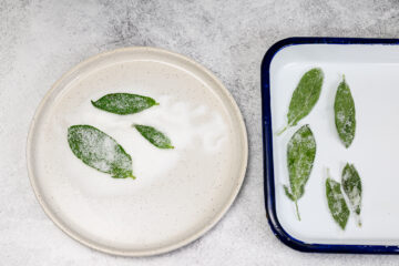 Green leaves coated in sugar on a white plate and a tray, set on a light gray surface.