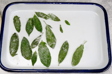 Green leaves coated with sugar are arranged on a white enamel tray with a blue rim.
