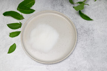 A round plate with a pile of white powder, likely citric acid, surrounded by green leaves on a light gray surface.