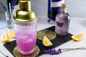 A glass cocktail shaker with a gold lid is filled with a purple drink, surrounded by lemon wedges, a bottle of purple liquid, and sprigs of lavender on a marble surface.