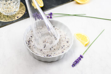 A glass grinder crushing lavender salt in a white bowl, with lemon wedges and lavender sprigs on a white surface.