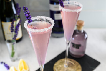 Two pink cocktails in flute glasses with sugar-rimmed edges and lavender sprigs as garnish, surrounded by bottles and lemon wedges on a marble surface.