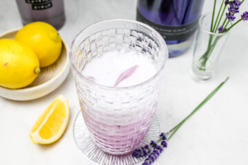 A glass of purple fizzy drink with ice sits on a coaster, surrounded by lemons, a bottle, a lavender sprig, and a glass with more lavender.