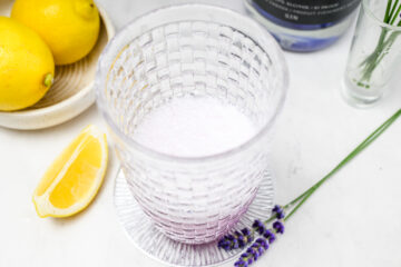A glass with a light purple foamy drink sits on a white surface, next to lemons, a lemon wedge, and sprigs of lavender.