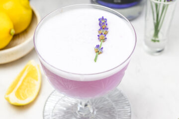 A coupe glass filled with a frothy lavender-colored cocktail, garnished with a sprig of lavender; lemons are visible in the background.