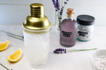 A cocktail shaker with a gold lid sits on a counter near lemon wedges, lavender sprigs, a bottle labeled lavender syrup, and a jar of salt.