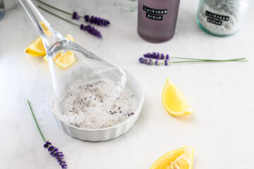 A cocktail glass is rimmed with lavender sugar in a shallow dish, surrounded by lemon wedges, fresh lavender, and bottles labeled lavender syrup on a white surface.