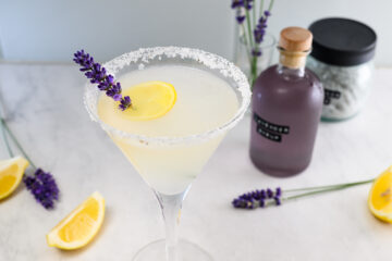 A cocktail in a martini glass with a salted rim, garnished with a lemon slice and lavender sprig, surrounded by lemons, lavender, and a jar of purple liquid.