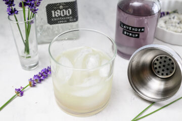 A glass of cocktail with ice sits on a white surface, surrounded by a bottle labeled Lavender Syrup, a tequila bottle, lavender flowers, and a cocktail shaker.