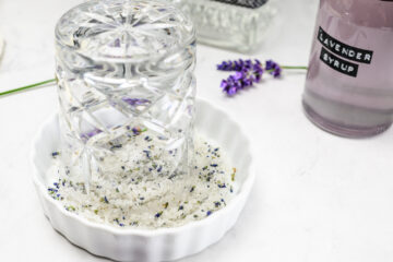 A glass presses into a dish of salt mixed with lavender buds, with a bottle labeled Lavender Syrup and a sprig of lavender nearby on a white surface.