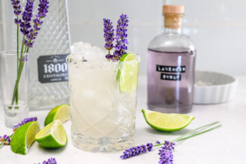 A glass filled with a lavender cocktail garnished with lavender sprigs and a lime wedge, with a bottle of lavender syrup, lime slices, and a tequila bottle nearby.
