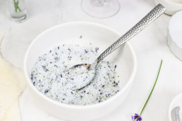 A white bowl filled with a coarse salt mixture and small purple flower pieces, with a metal spoon resting inside.