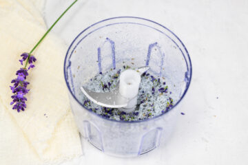 A food processor bowl with a mixture of salt and lavender buds inside, next to a cloth and a sprig of fresh lavender on a white surface.