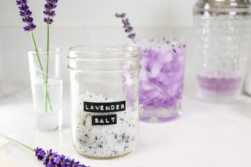 A glass jar labeled Lavender Salt sits on a counter with lavender sprigs, a glass of purple drink with ice, and a cocktail shaker in the background.