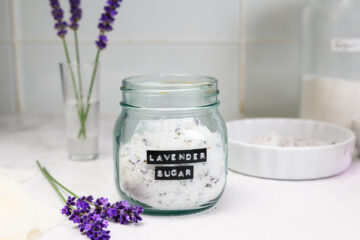 A glass jar labeled Lavender Sugar sits on a countertop, surrounded by fresh lavender sprigs and kitchen items.