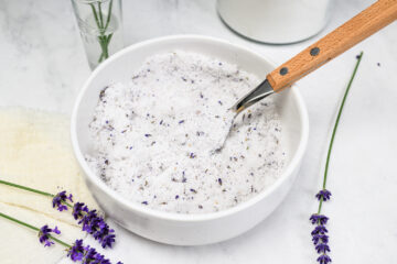 A white bowl filled with lavender-infused sugar, a metal spoon with a wooden handle, and fresh lavender sprigs beside it on a marble surface.
