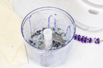 A food processor bowl containing a mixture of sugar and lavender buds, with a sprig of lavender and a cloth nearby on a white surface.