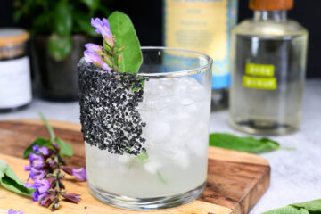 A cocktail with ice in a glass, rimmed with black salt and garnished with a green sage leaf and purple sage flowers, sits on a wooden board with a bottle of mezcal in the background.