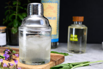 A metal cocktail shaker filled with ice sits on a wooden board, with bottles and herbs in the background.