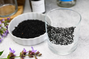 A clear glass with a section coated in black salt sits beside a white dish filled with black salt, with purple sage flowers nearby on a light surface.