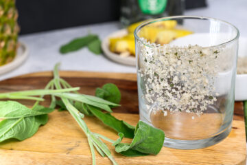 A glass rimmed with coarse salt sits on a wooden board next to fresh sage leaves, with pineapple slices and a bottle in the background.