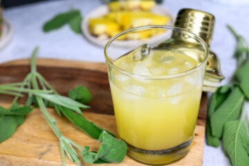 A glass of yellow cocktail with ice sits on a wooden board, surrounded by fresh green green leaves and a gold cocktail shaker in the background.
