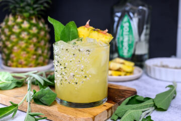 A glass of yellow cocktail with ice, pineapple garnish, and salt-rimmed edge sits on a wooden board with sage leaves, a pineapple, and a bottle in the background.