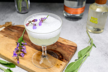 A cocktail in a coupe glass topped with foam and purple flowers, placed on a wooden board with sage leaves and bottles in the background.