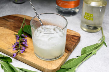 A glass of iced cocktail is being stirred on a wooden board with fresh sage leaves and purple flowers nearby; bottles and sage syrup are in the background.