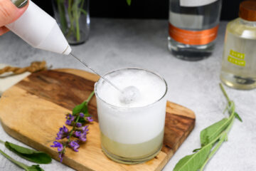 A hand uses a handheld frother to foam the top of a cocktail in a glass on a wooden board, with sage, a bottle, and a jar nearby.
