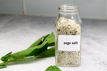 A glass jar labeled sage salt is filled with a coarse salt and herb mixture, with fresh sage leaves lying next to it on a light surface.