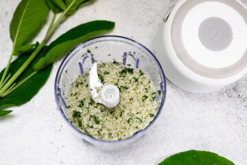 A food processor bowl filled with chopped herbs and coarse mixture, surrounded by fresh green sage leaves on a white surface.