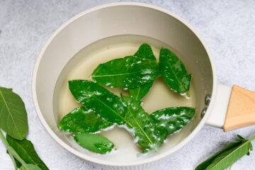 A white saucepan filled with sugar water and several fresh green sage leaves, placed on a light gray surface.