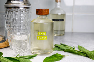 A glass bottle labeled Sage Syrup stands beside a shot glass, cocktail shaker, and sage leaves on a countertop.