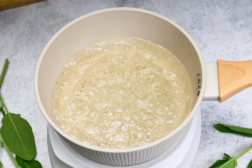 A white saucepan with a wooden handle filled with water and sugar at a rolling boil on a white stovetop, with sage herb leaves nearby.