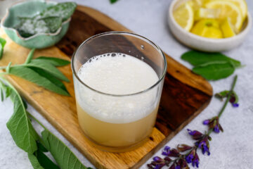 A glass of frothy light-colored beverage sits on a wooden board, surrounded by lemon slices, fresh sage leaves, and purple flowers.