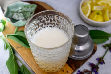 A glass filled with a frothy light-colored drink sits on a wooden board next to a metal shaker, fresh herbs, and a bowl of lemon wedges.