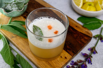 A frothy cocktail garnished with a sage leaf and three drops of bitters, served in a glass on a wooden board with fresh sage leaves and lemon wedges nearby.