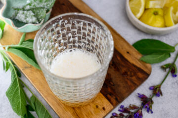 A glass with a small amount of frothy liquid sits on a wooden board, surrounded by sage leaves, purple flowers, and a bowl of lemon wedges.