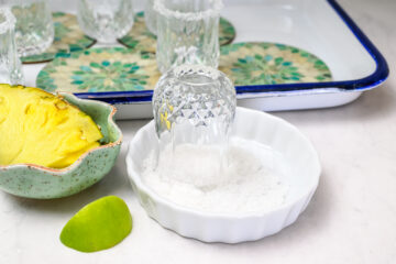 A glass is being rimmed with salt in a small white dish, with a pineapple slice, a lime wedge, and other glasses on a tray in the background.