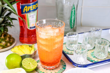 A glass of orange cocktail with ice sits on a counter next to a lime, pineapple slices, Aperol bottle, Hornitos tequila bottle, and several empty shot glasses.