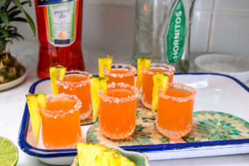 Six orange cocktails with salted rims and pineapple wedges are arranged on a tray, with bottles and a sliced lime in the background.